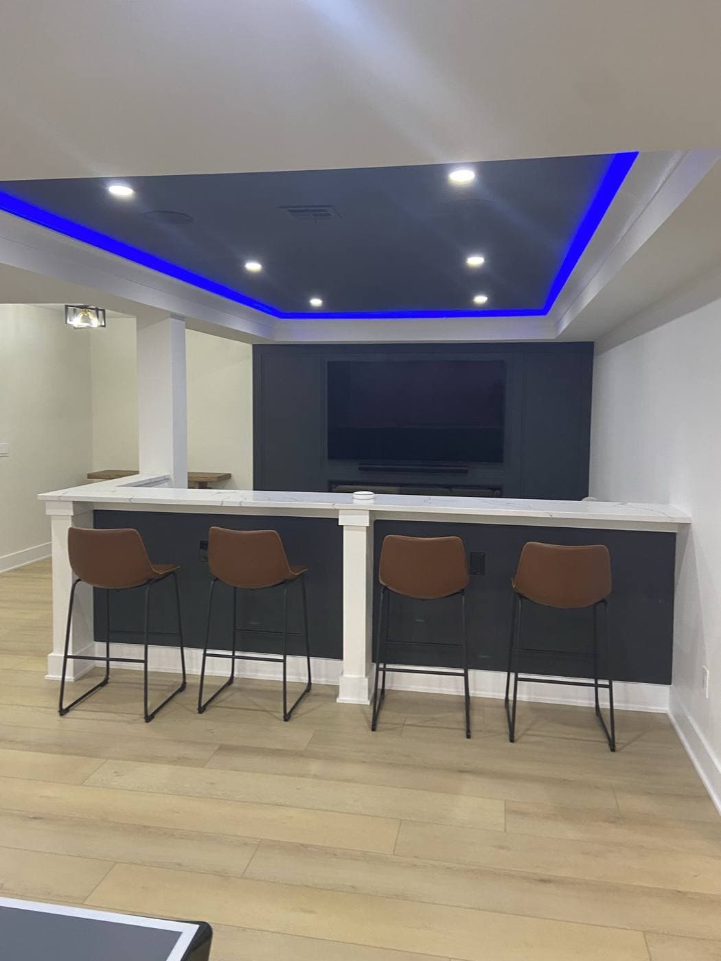 A basement bar and TV wall with blue cove lighting tracing the full perimeter of a tray ceiling and recessed cans inside it.