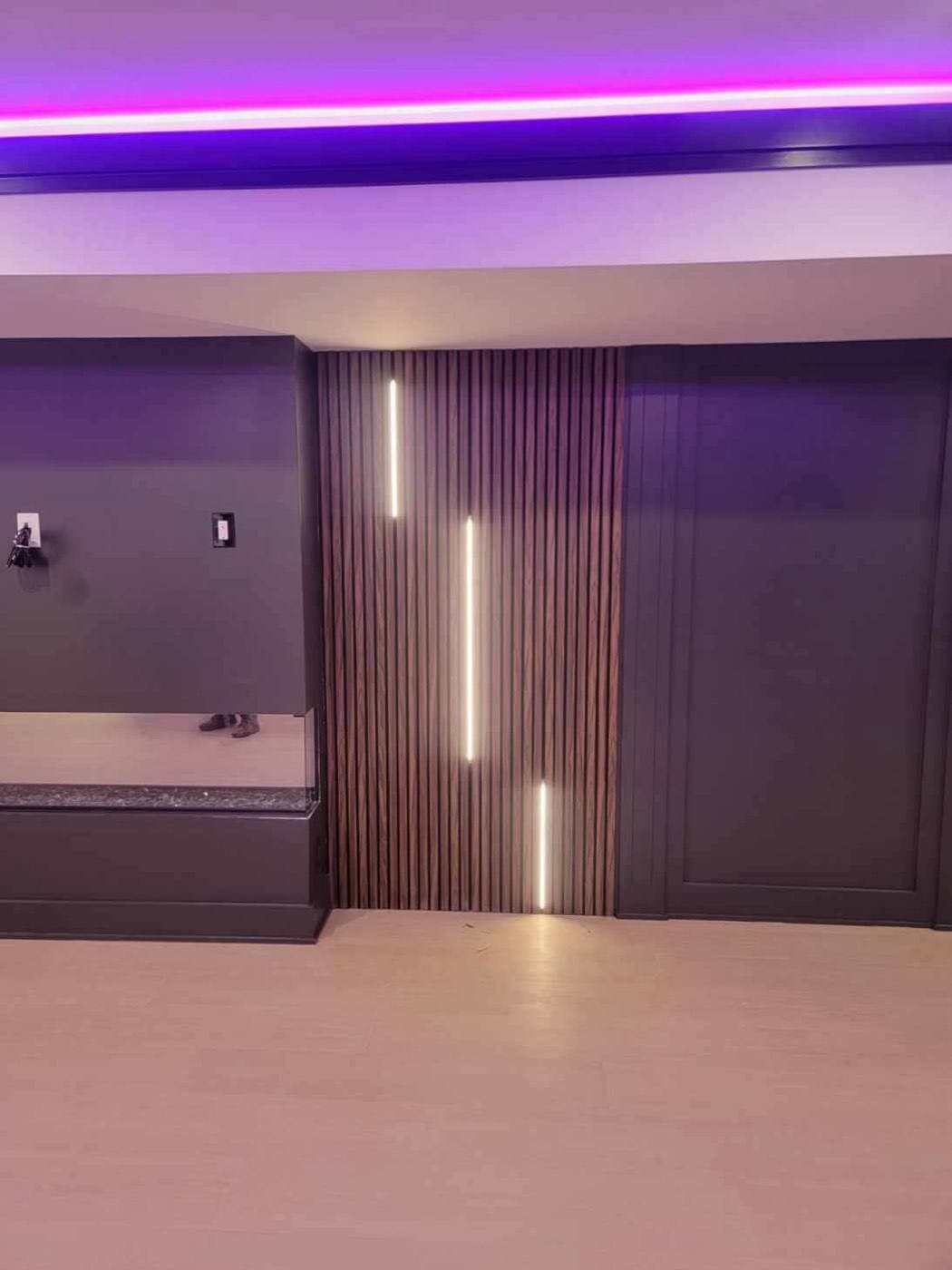 A wood slat wall with three vertical linear light fixtures at staggered heights, purple cove lighting overhead.